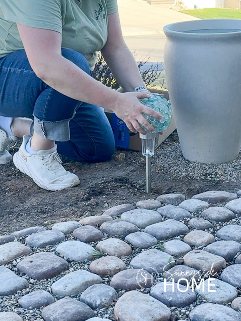 Woman places glass gem orb over dollar store solar lights along the edge of a cobble stone path.