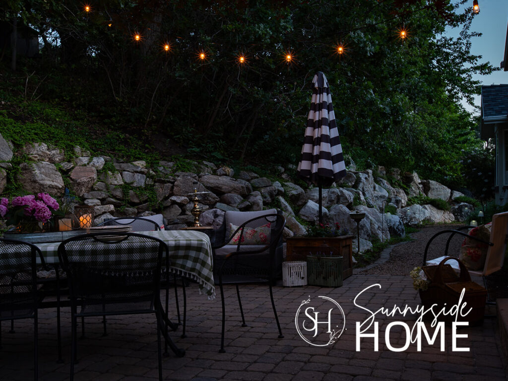 Dusk the string lights over the styled patio with a charming cottage vintage vibe using floral and gingham fabric .