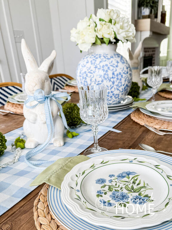 Blue and white Easter table settings with a blue and white vase with peonies centerpiece on a blue and white gingham table runner embellished with bunnies and moss balls, water hyacinth placemats, blue and white striped plate, sage green napkin and a floral plate.