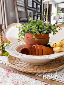 A collection of thrift store clay pots are styled in a thrifted bowl with greenery and tulips on a dining table with brass candlesticks.