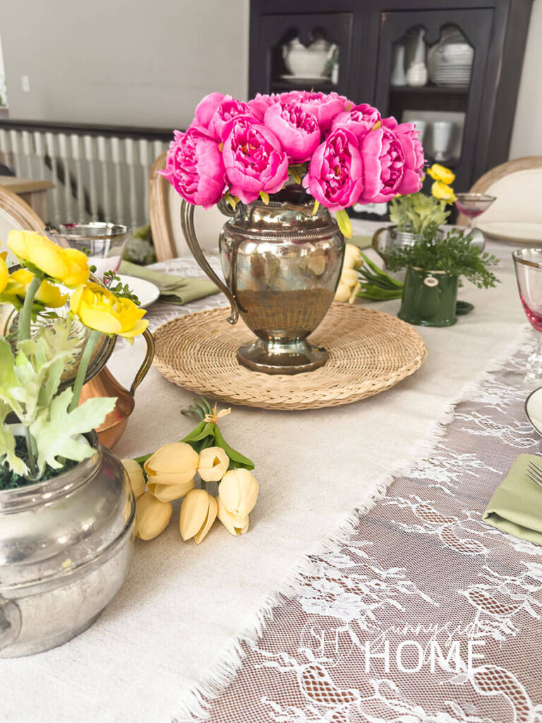 Easter centerpiece, a linen table runner is placed over a lace tablecloth, a silverplate pitcher is filled with pink peonies on a wicker tray and additional greenery and yellow flowers in small teapots.