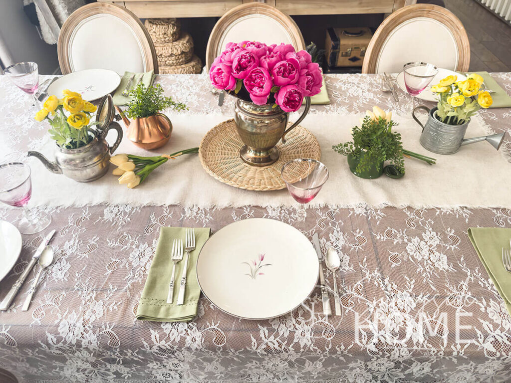 Easter tablescape, vintage pink lily plate, sage green napkin, pink hue goblets and vintage silverware. For the centerpiece a linen table runner is placed over a lace tablecloth, a silverplate pitcher is filled with pink peonies on a wicker tray and additional greenery in small teapots.