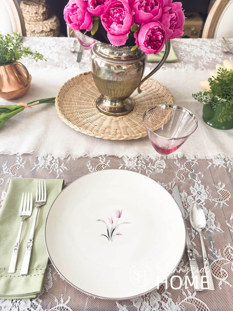 Easter table settings, vintage pink lily plate, sage green napkin, pink hue goblets and vintage silverware. For the centerpiece a linen table runner is placed over a lace tablecloth, a silverplate pitcher is filled with pink peonies on a wicker tray and additional greenery in small teapots.