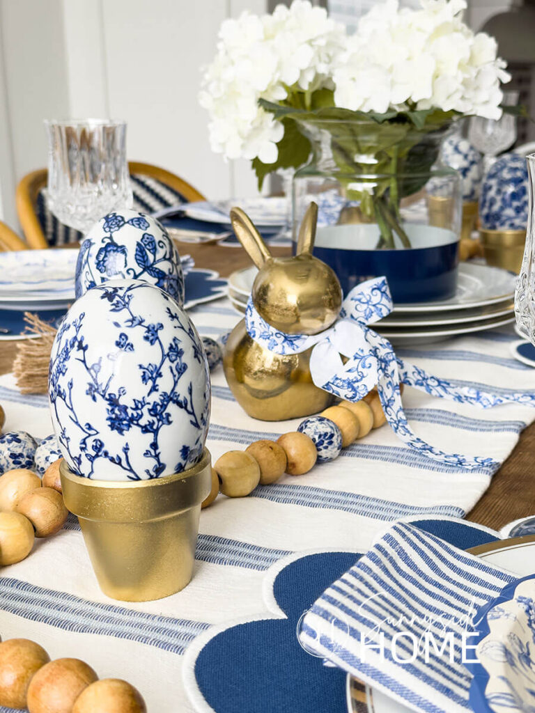 Easter table centerpiece is a blue and white table runner, white hydrangeas in a blue and glass color blocked vase, gold bunnies, chinoiserie eggs and a wood bead garland.