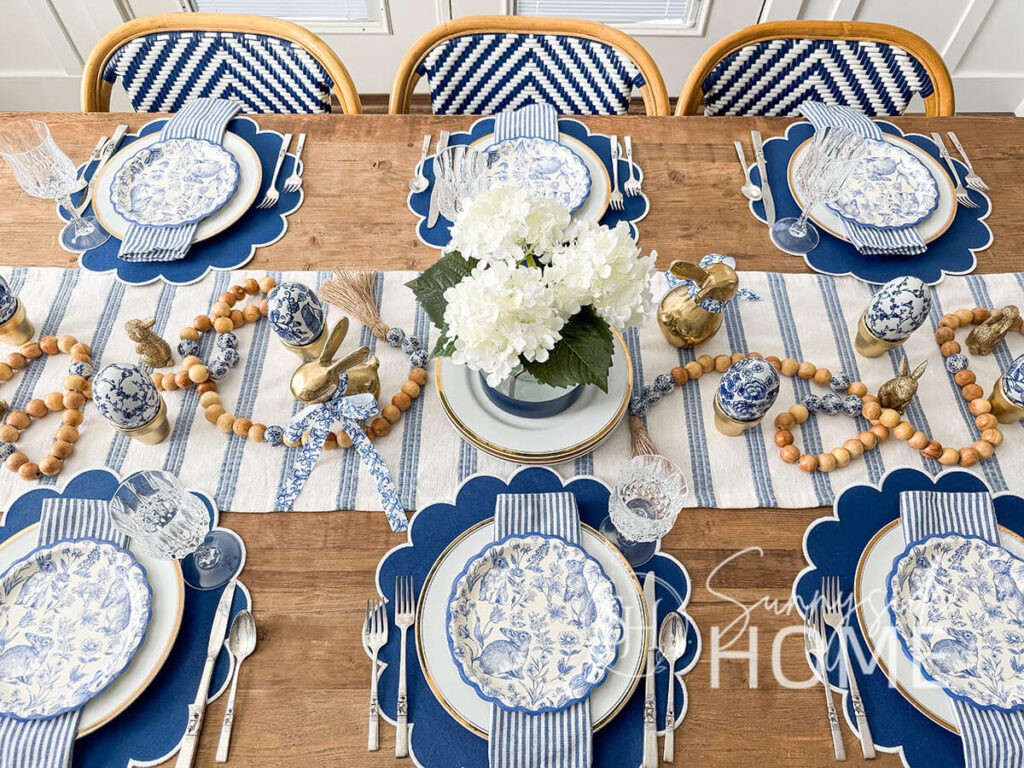 Easter tablescape in shade of blue and white, at each place setting is Blue scalloped place mat topped with a gold rimmed plate, blue and white striped napkin and blue and white bunny and botanical scalloped paper plate. For the centerpiece there is a blue and white table runner, white hydrangeas in a blue and glass color blocked vase, gold bunnies, chinoiserie eggs and a wood bead garland.