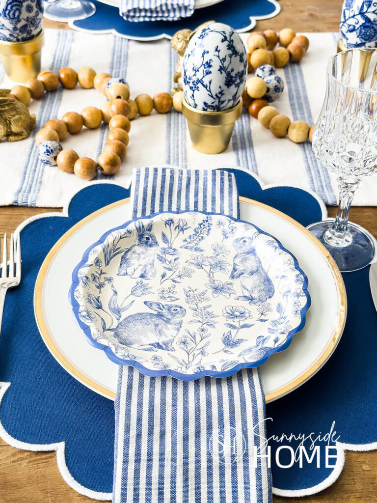 Easter table settings: Blue scalloped place mat topped with a gold rimmed plate, blue and white striped napkin and blue and white bunny and botanical scalloped paper plate.