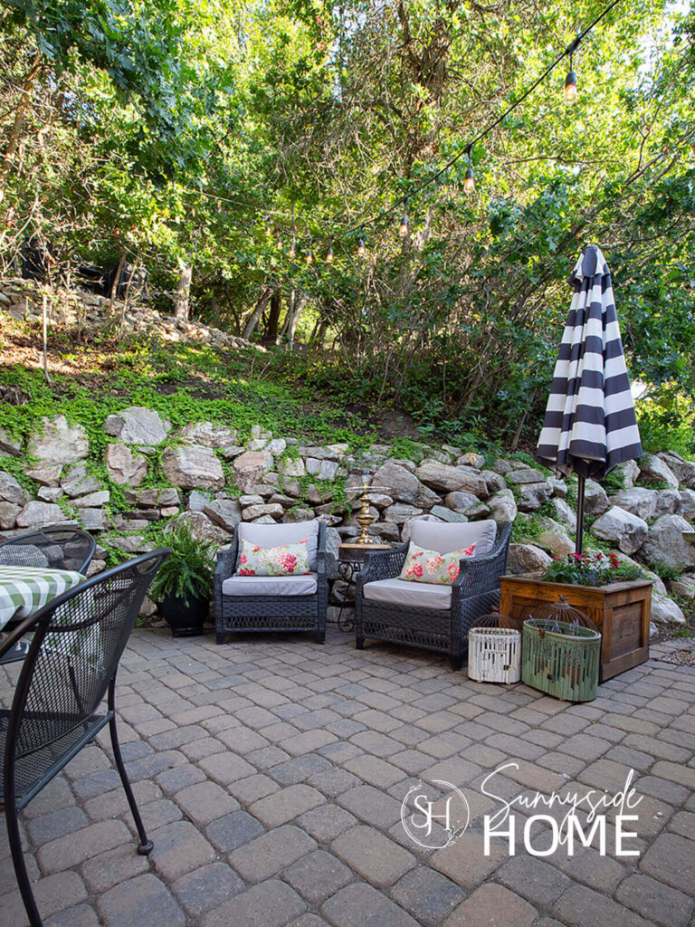 Seating area on the patio with a pair of wicker chairs with cushion and floral pillowd, potted fern, planter box with dahlias and a black and white striped umbrella.