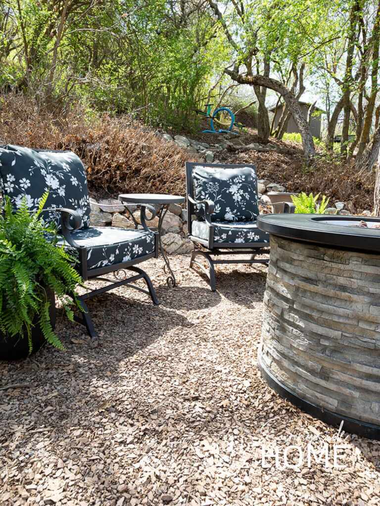 Patio chairs around a fire pit in a wooded backyard.