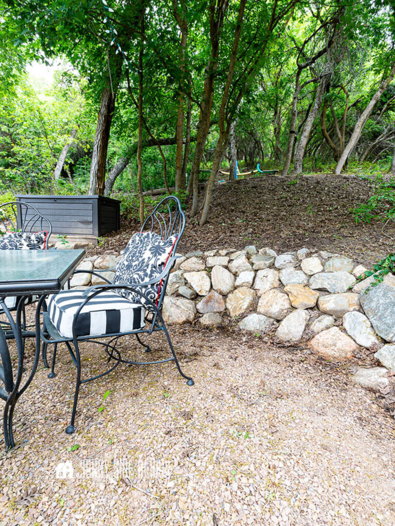 A simply rock rock retain wall on a wooded backyard landscape with a dining table and deck box.