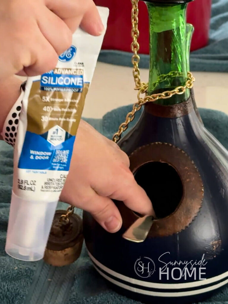Woman holds a tube of silicone to attach an old metal spoon to a painted blue wine decanter to create a birdhouse perch.