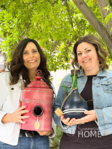 Two women hold their whimsical DIY birdhouses made from thrifted finds, a red canister and a wine decanter.