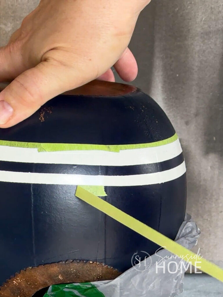 Woman removes green painters tape to reveal white stripes painted on the navy blue wine decanter birdhouse.