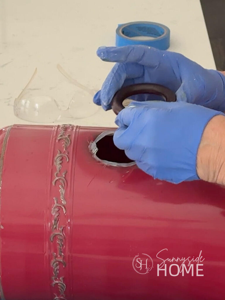 Woman attaches a round wooden drapery ring to the hole of the red canister birdhouse with outdoor silicone adhesive.