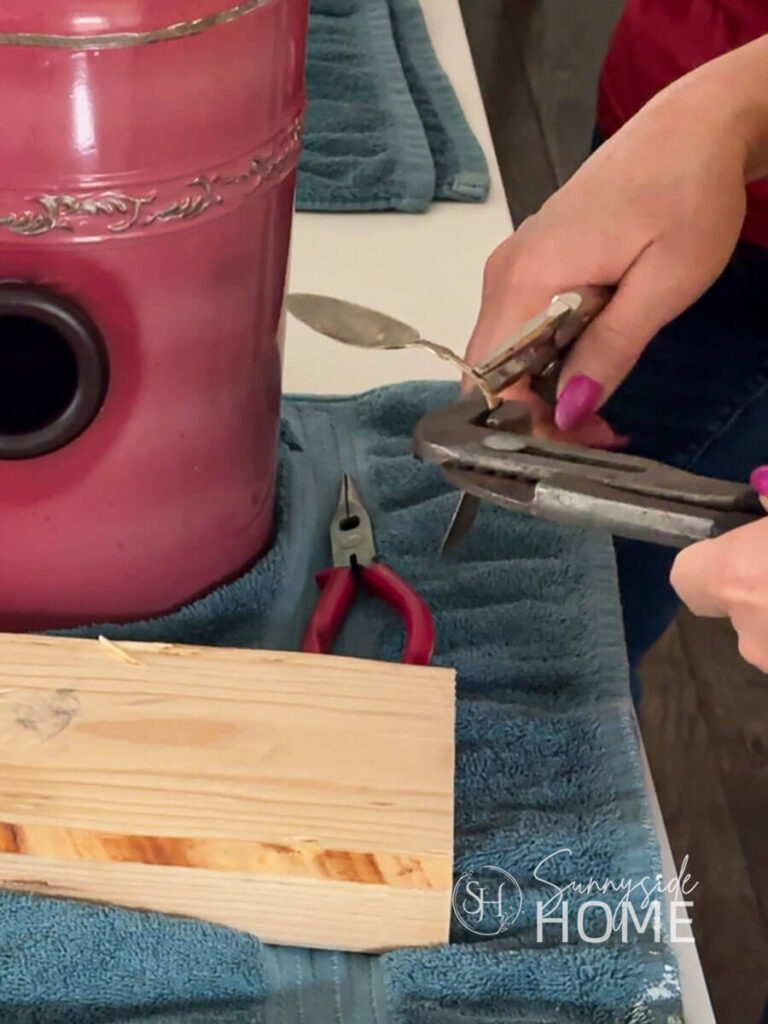 Woman uses pliers to bend an old metal spoon to create a perch for the red metal canister birdhouse.