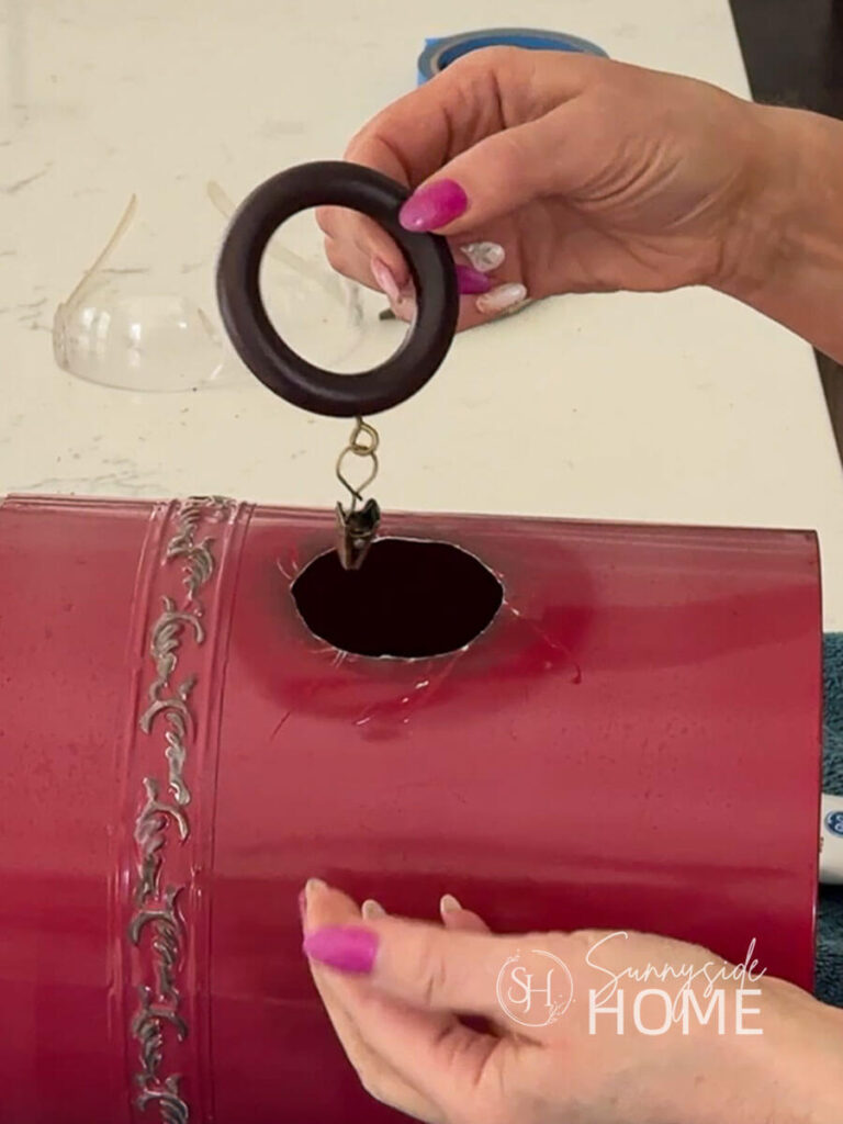 Woman holds a wooden drapery ring at opening on red metal canister birdhouse.