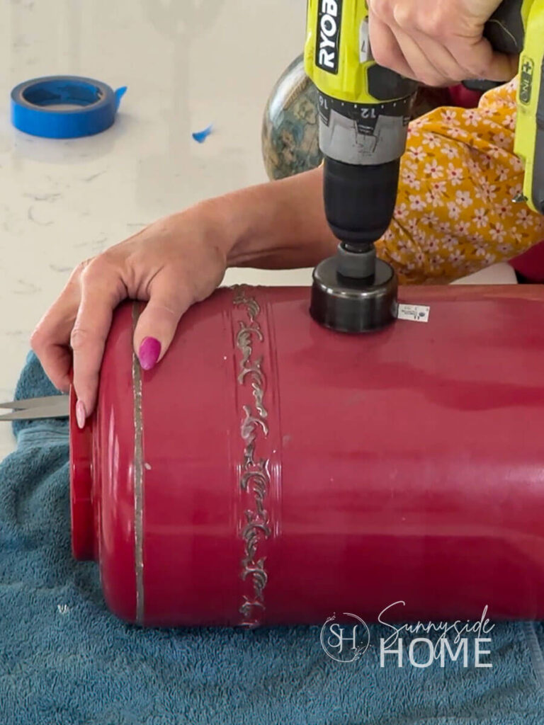 Woman uses a drill with a hole saw to drill a hole in a red metal canister for a clever garden birdhouse.