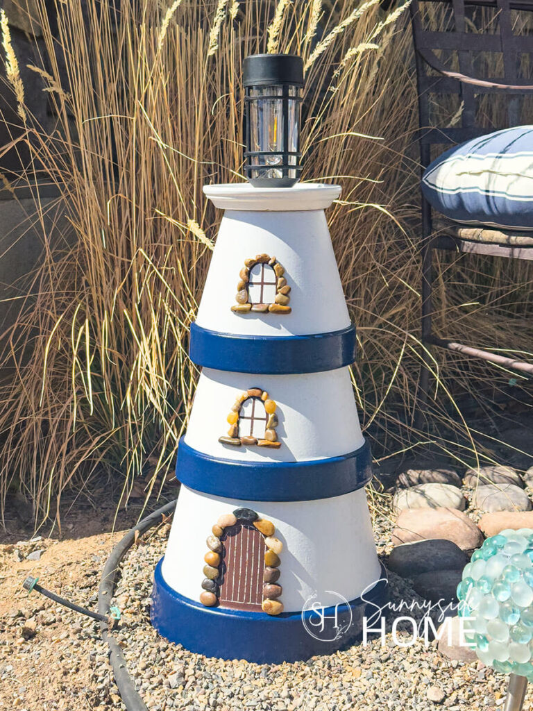 Simple DIY clay pot lighthouse, white with navy blue bands and a solar light stop, embellished with pebbles around the door and windows, styled in a garden with grasses.