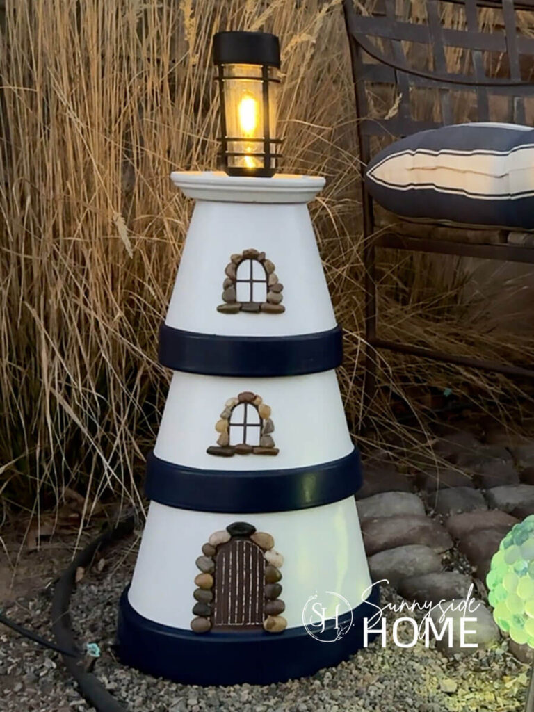 Simple DIY clay pot lighthouse, white with navy blue bands and a solar light stop, embellished with pebbles around the door and windows, styled in a garden with grasses.