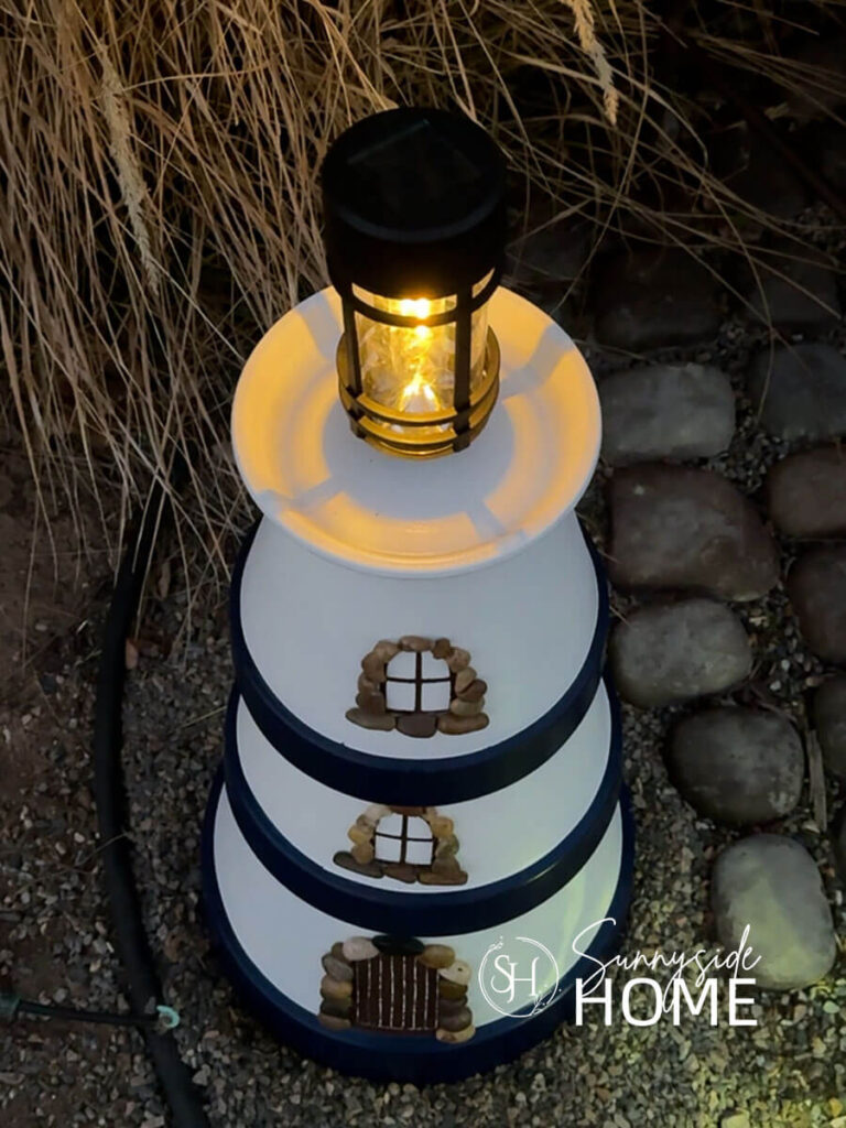 Simple DIY clay pot lighthouse, white with navy blue bands and a solar light stop, embellished with pebbles around the door and windows, styled in a garden with grasses.