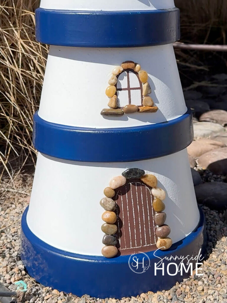 Simple DIY clay pot lighthouse, white with navy blue bands and a solar light stop, embellished with pebbles around the door and windows, styled in a garden with grasses.