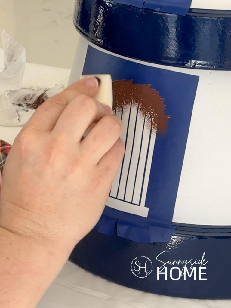 Woman uses a cosmetic sponge to apply brown paint to the door stencil on the pained clay pot lighthouse.
