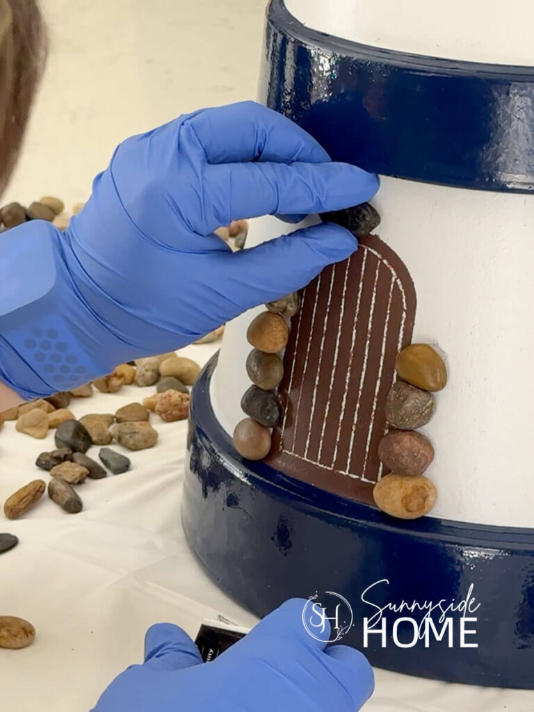Woman adheres pebbles with silicone around the stenciled door on the clay pot lighthouse.