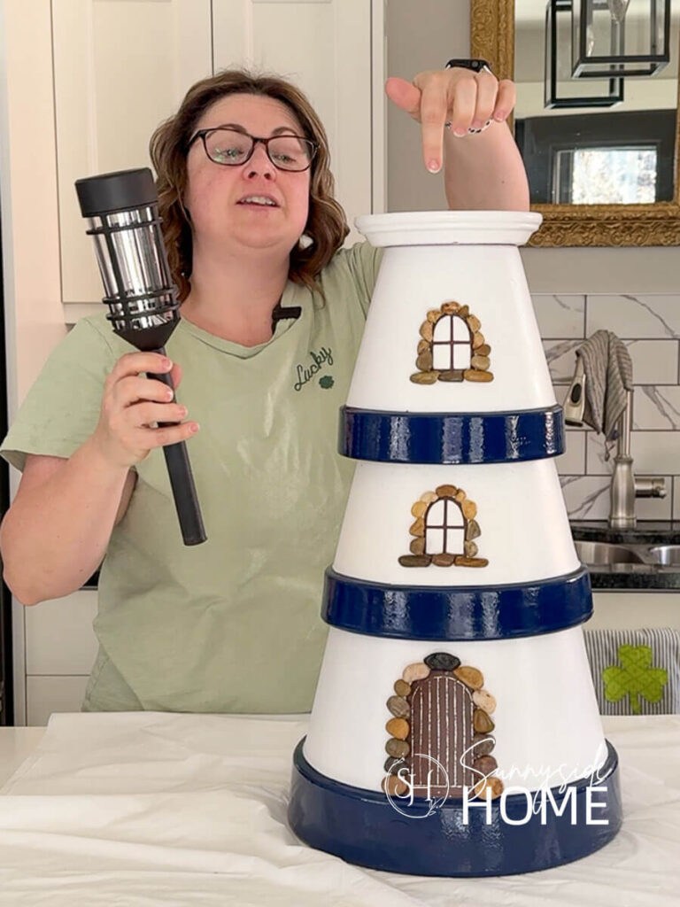 Woman holds a solar light and points to the hole in the top of the clay pot lighthouse garden decor.