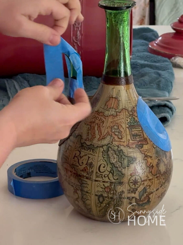 Woman applies blue painters tape to a thrifted wine decanter to prepare if for paint.