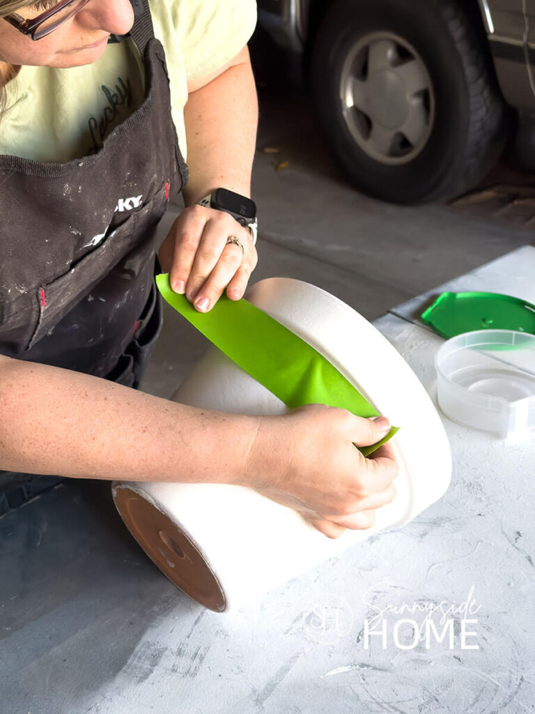 Woman applies painters tape below the rim of the white painted clay pot.