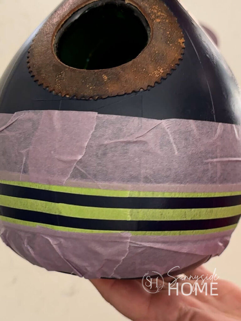 Woman holds painted blue decanter birdhouse to show placement of painters tape to make stripes on her decanter birdhouse.
