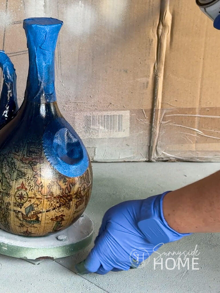 Woman applies navy blue spray paint to the wine decanter birdhouse.