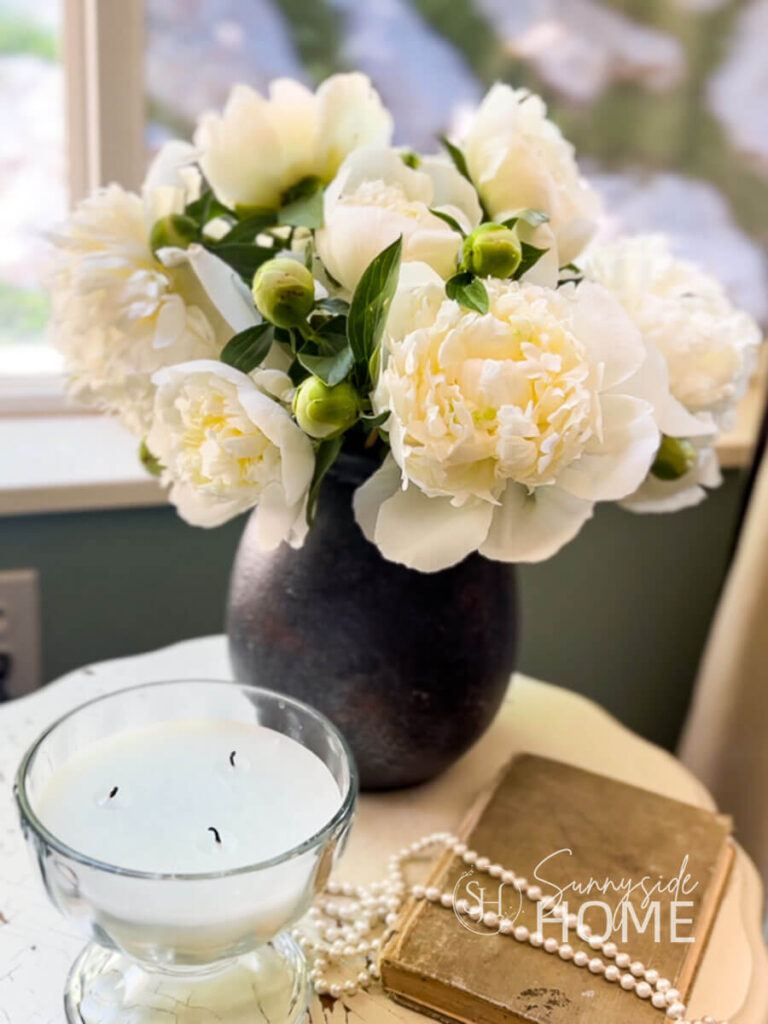 White peonies arranged in a white vase, styled on an accent table with a candle, book and a strand of pearls.