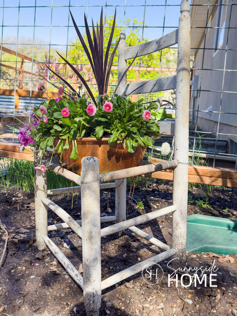 A thrifted copper pot is turned into a planter container and filled with assorted flowers and dracena and placed on an old chippy wood chair.