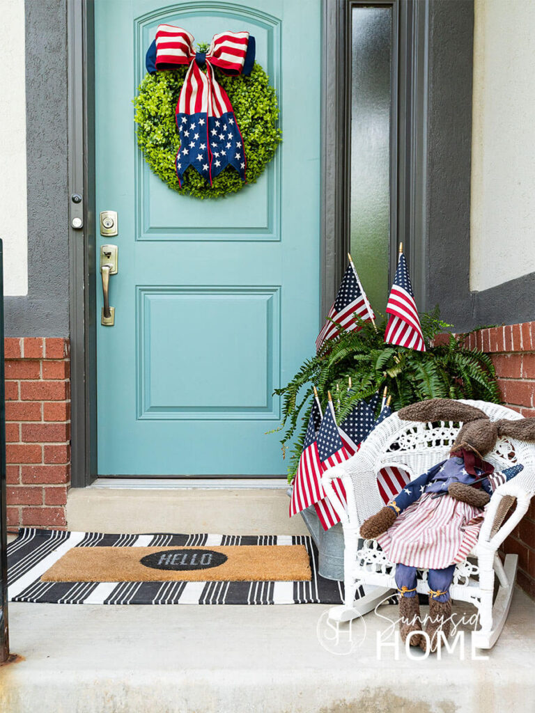 Decorating ideas for summer on the front porch. A boxwood wreath is hung on a blue door and embellished with a patriotic red, white and blue bow. A potted fern with 2 American flags, galvanized vintage watering can filled with more American flags and a white child wicker rocker is styled with a patriotic dressed bunny with floppy legs.