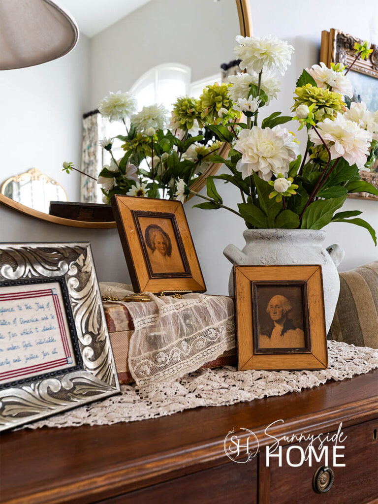 The entryway is decorated for summer with vintage lace on a child's suitcase with vintage framed images of George and Martha Washington, framed hand stitched pledge of allegiance.