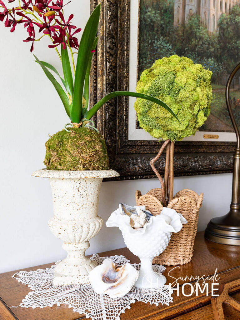 Simple summer decorations on the piano, a rustic vase with an orchid, a scalloped basket with a moss topiary, milk glass footed bowl filled with seashells.