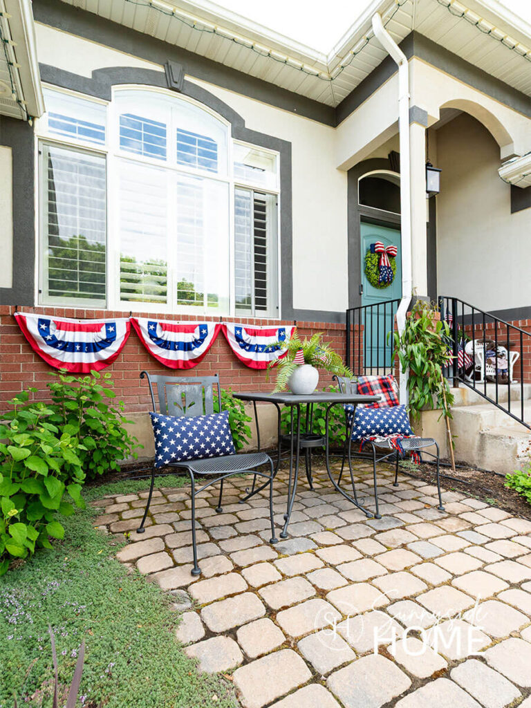 Outdoor Decorating ideas for summer. A cobblestone patio with a bistro table styled with blue star pillows and a red, white and blue plaid throw. A patriotic bunting is hung below the window and a boxwood wreath embellished with a patriotic bow is hung on the blue front door.