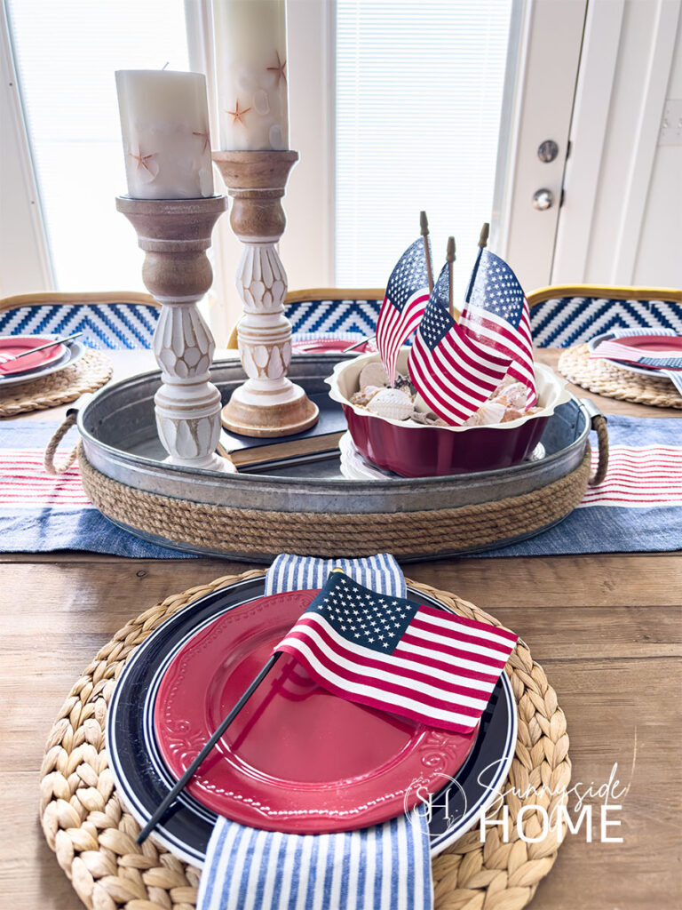 Dining table set for a 4th of July celebration with water hyacinth placements topped with a striped blue place, striped chambray napkin, red plate and a small American flag. The centerpiece consists of a pair of coastal candles in wood candlesticks and a red bundt ban filled with sand and seashells with 3 small American flags in the center, styled in an oval galvanized tray. The tray is placed on a red, white and blue tablerunner.