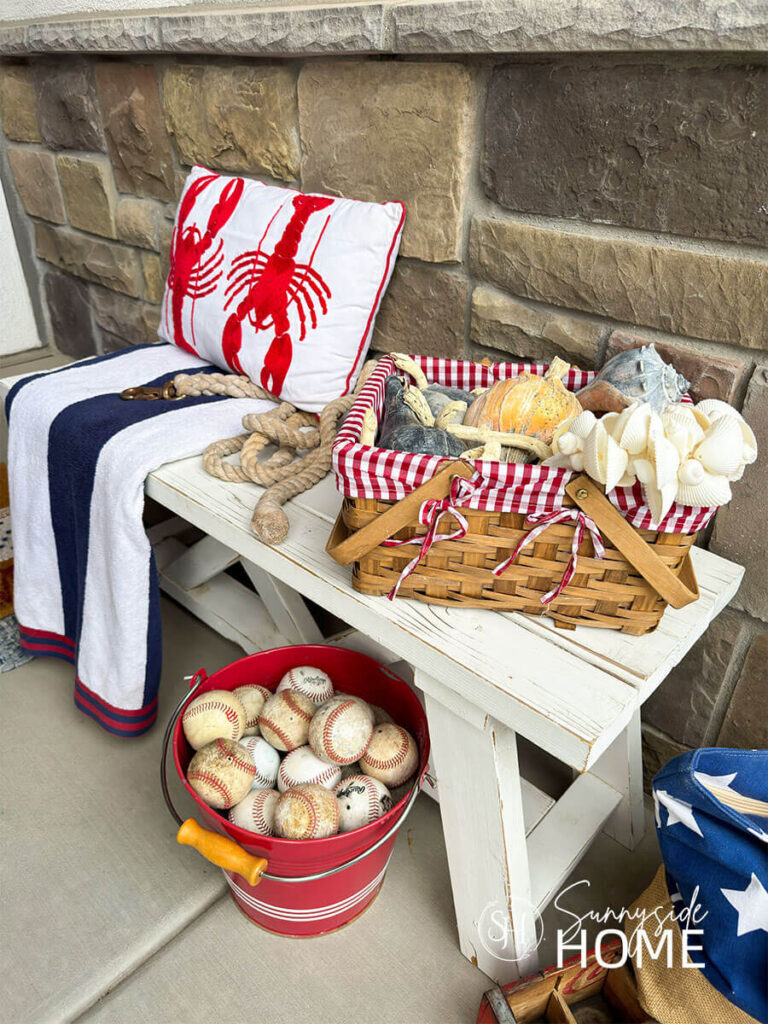 Fourth of July decorating ideas for the front porch, a white farmhouse bench is styled with a red, white and blue beach towel, red lobster pillow, basket with a red and white gingham liner filled with seashells, under the bench if a red bucket filled with baseballs.
