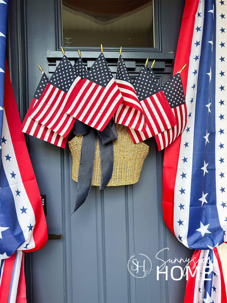 A basket filled with American flags and embellished with a blue denim bow is hung on a navy blue front door. A patriotic bunting is hung around the door frame.
