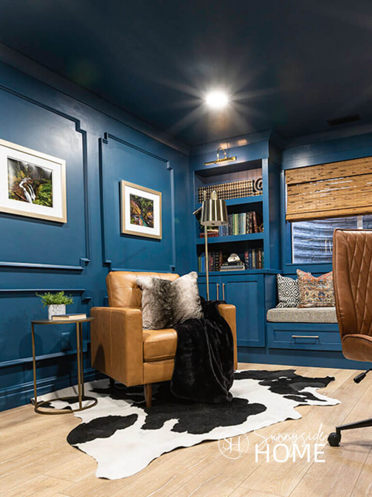 A home office with build in bookshelves and library lights are painted a moody blue with a cozy leather club chair, floor lamp, side table, faux fur pillow and throw, styled with a cow hide rug.