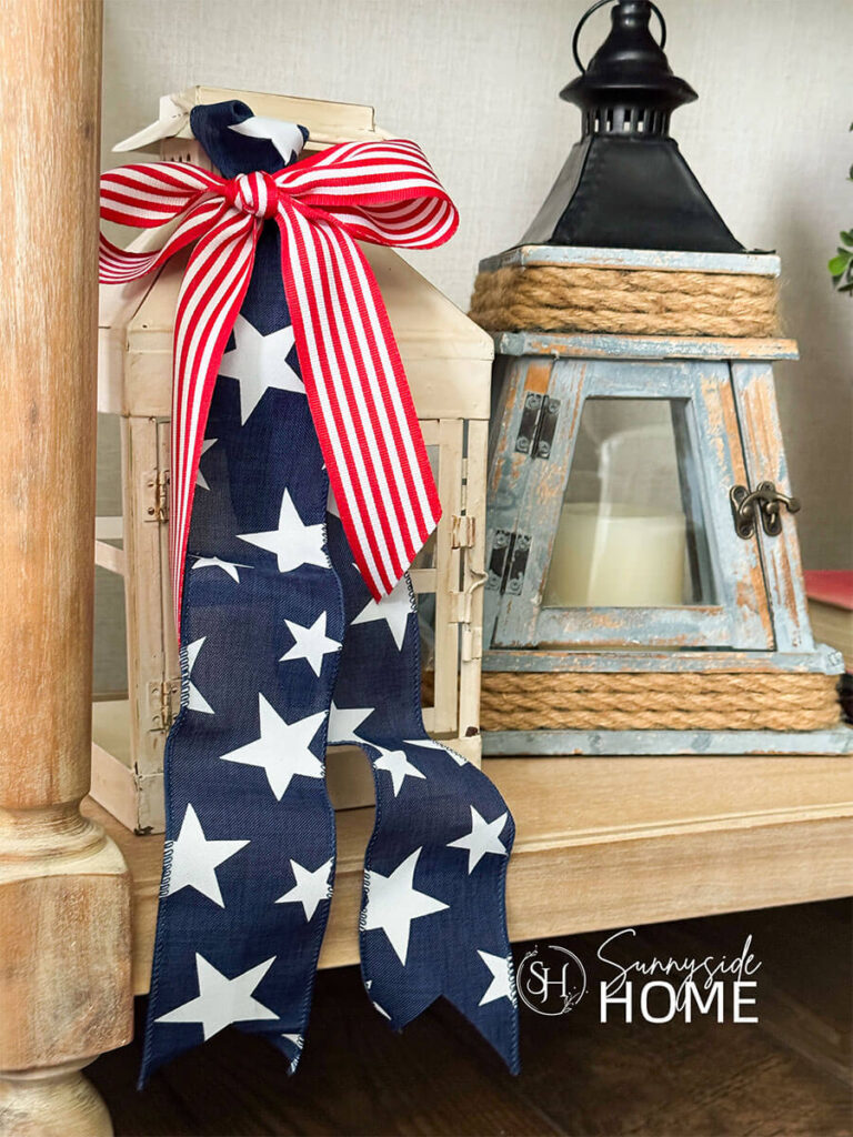 On the lower shelf of the sofa table is styled with lanterns, one is embellished with a blue ribbon with white stars and tied with a red and white bow.