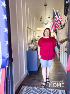 Woman in red shirt and jeans opening door to her fourth of July decorations in her home. Basket of flags on a navy blue door, patriotic bunting around door, blue entryway cabinet decorated with summer patriotic decor.