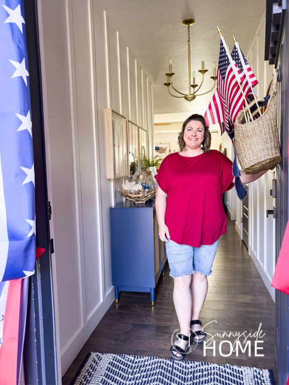 Woman in red shirt and jeans opening door to her fourth of July decorations in her home. Basket of flags on a navy blue door, patriotic bunting around door, blue entryway cabinet decorated with summer patriotic decor.