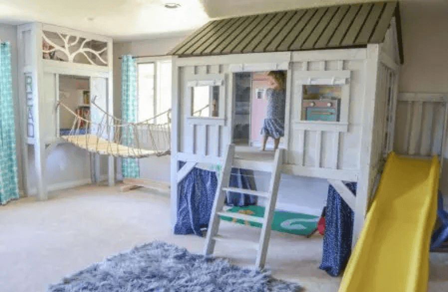 In a child's playroom a cozy treehouse reading nook is created against the wall and is connected to a elevated playhouse with a suspension bridge.