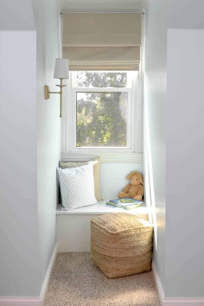 A dormer window with a built in bench, pillows, wall sconce, books and ottoman.