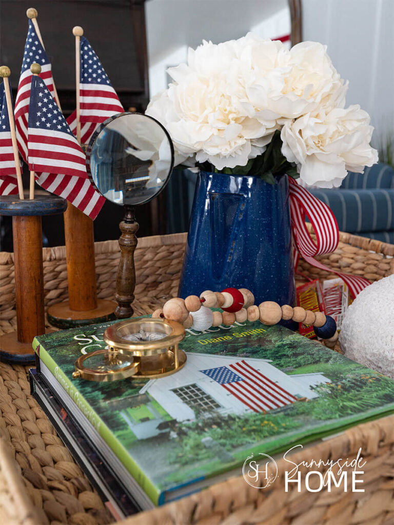 Fourth of July decorating ideas in a basket tray on the coffee table: a stack of summer travel books, spools with flags, blue coffee pot filled with white peonies and embellished with red and white striped ribbon.