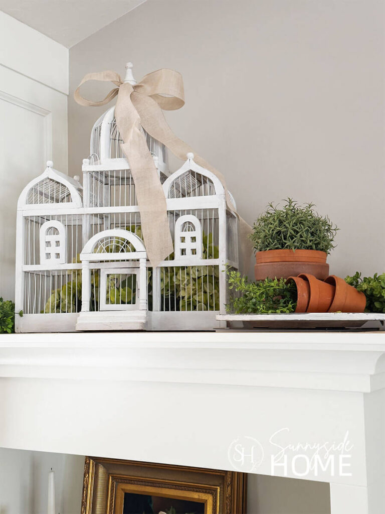 Decorating the top of a built in bookcase with a vintage white birdcage filled with light green hydrangeas and a linen bow. Also in the vignette are stacked clay pots and greenery.