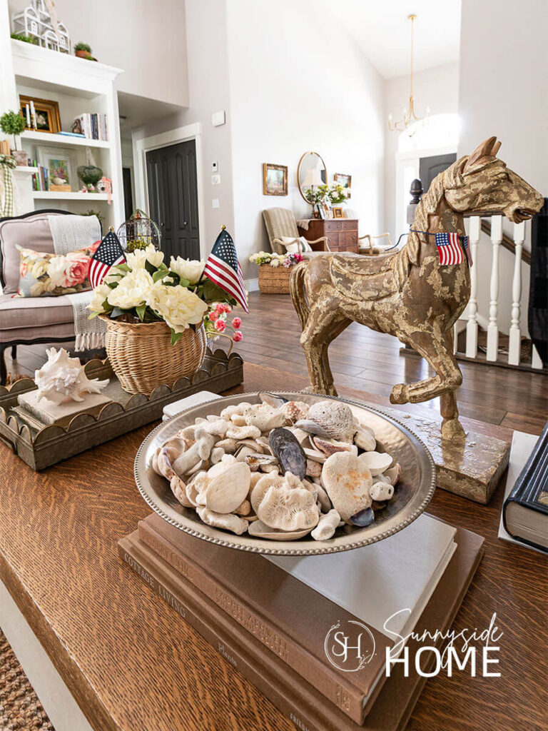 Decorating ideas for summer on the coffee table. a pewter footed bowl is filled with collected seashells, a wood horse statue is embellished with a mini flag bunting, a scalloped galvanized tray is filled with a wicker base with white peonies and 2 American flags and a large seashell sits a top a vintage book stack.