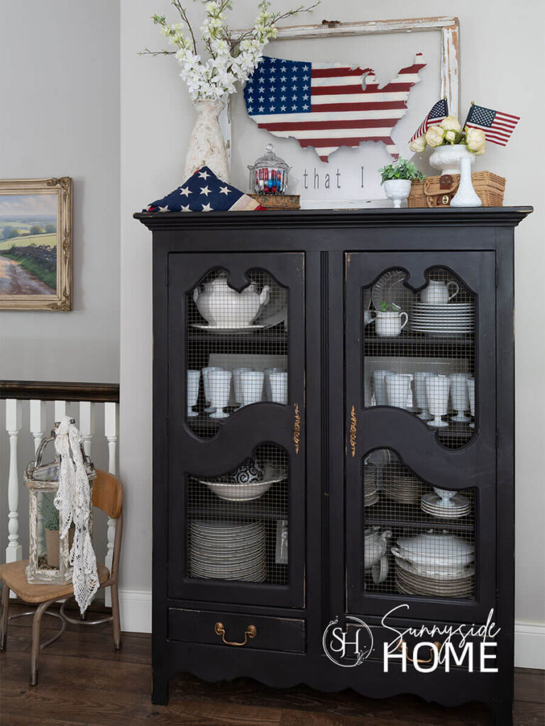 A black china cabinet is decorated for summer with a hand painted vintage window with a patriotic scene, white florals, milk glass and textured vases, folded American flag, crystal container filled with patriotic gumballs.